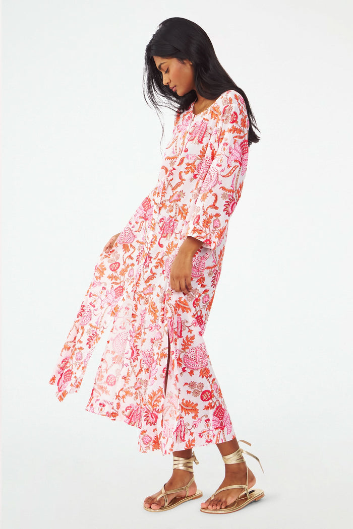 A woman with long dark hair wears the Amanda Anu Long Kurta by Roller Rabbit, a flowy paisley print dress with long sleeves. She stands sideways in gold strappy sandals against a plain white background, looking down.