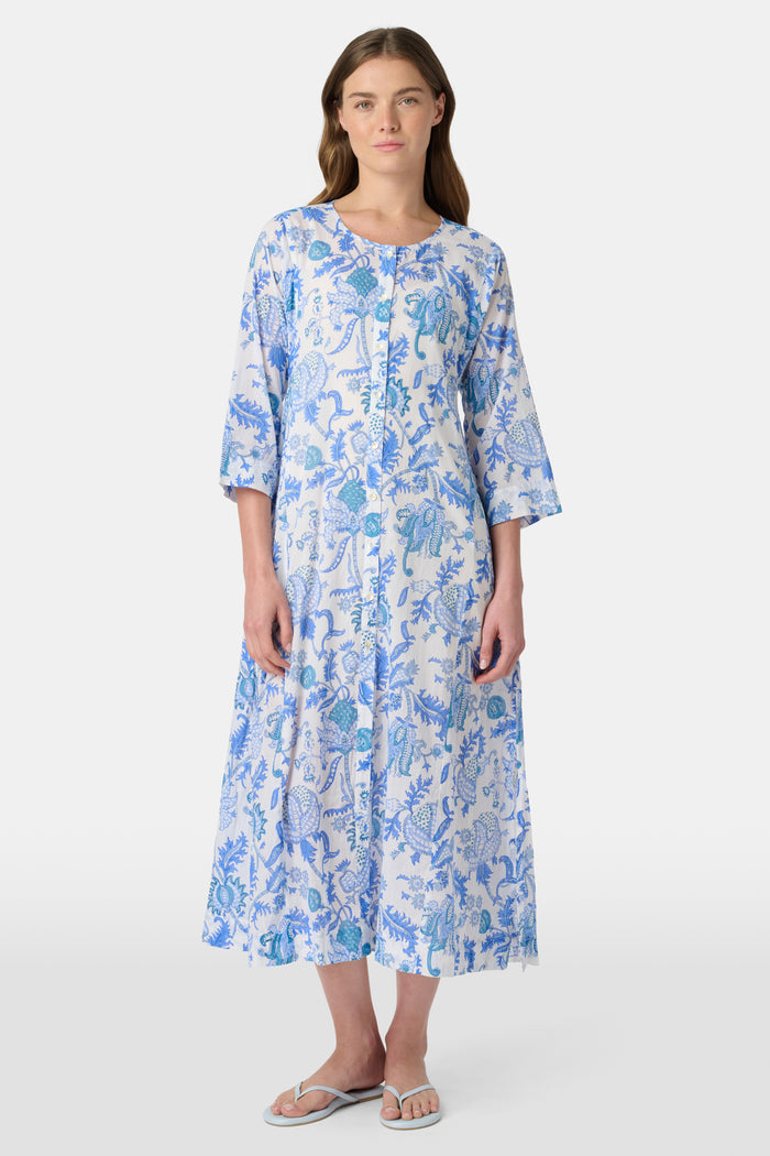 A woman faces forward against a plain white background, wearing the Amanda Anu Long Kurta by Roller Rabbit in blue and white floral cotton voile with three-quarter sleeves, paired with white flip-flops.