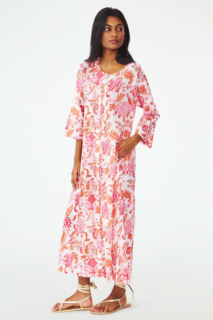 A woman with long dark hair wears the Amanda Anu Long Kurta by Roller Rabbit, a flowy paisley print dress with long sleeves. She stands sideways in gold strappy sandals against a plain white background, looking down.