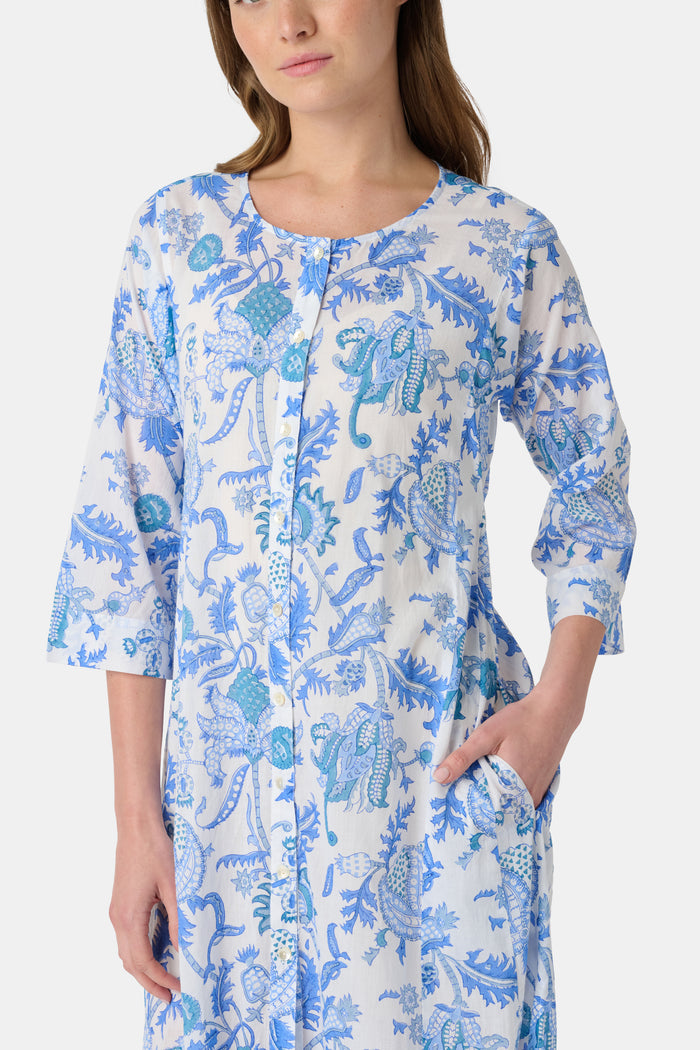A woman faces forward against a plain white background, wearing the Amanda Anu Long Kurta by Roller Rabbit in blue and white floral cotton voile with three-quarter sleeves, paired with white flip-flops.