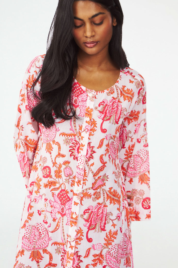A woman with long dark hair wears the Amanda Anu Long Kurta by Roller Rabbit, a flowy paisley print dress with long sleeves. She stands sideways in gold strappy sandals against a plain white background, looking down.