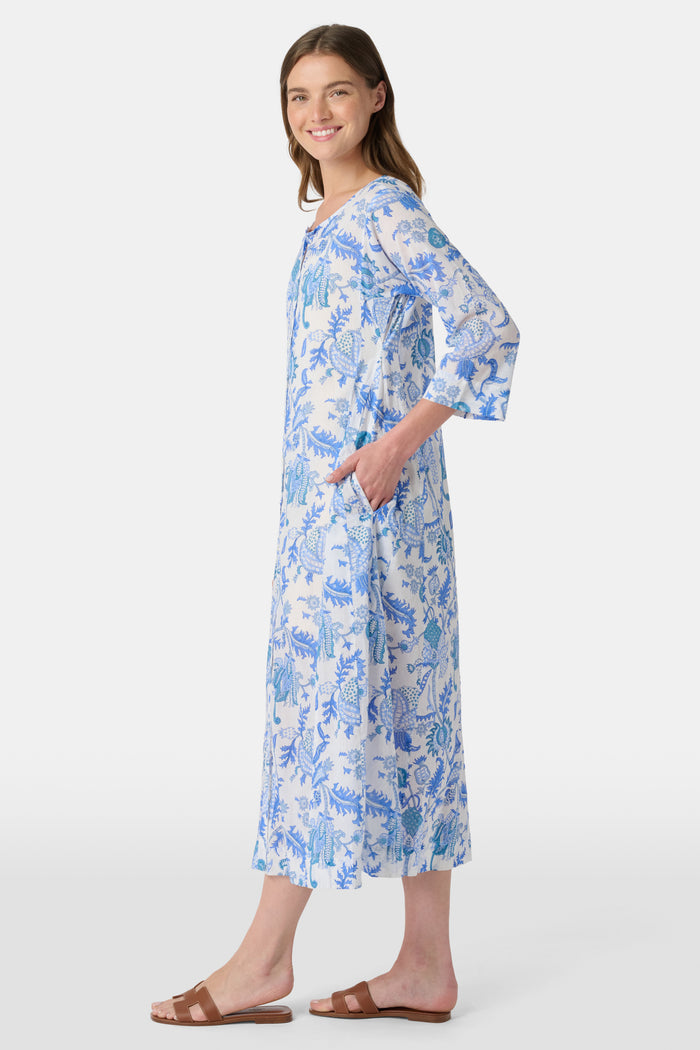 A woman faces forward against a plain white background, wearing the Amanda Anu Long Kurta by Roller Rabbit in blue and white floral cotton voile with three-quarter sleeves, paired with white flip-flops.