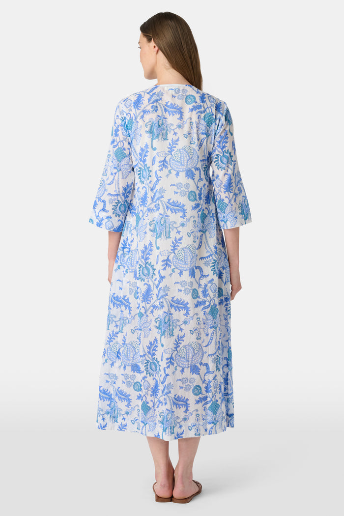 A woman faces forward against a plain white background, wearing the Amanda Anu Long Kurta by Roller Rabbit in blue and white floral cotton voile with three-quarter sleeves, paired with white flip-flops.