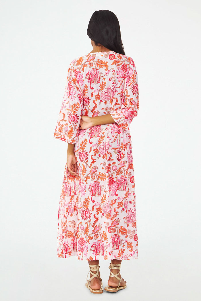 A woman with long dark hair wears the Amanda Anu Long Kurta by Roller Rabbit, a flowy paisley print dress with long sleeves. She stands sideways in gold strappy sandals against a plain white background, looking down.
