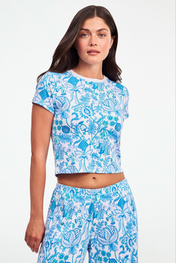 A woman with long brown hair wears the Roller Rabbit Amanda Baby Tee, featuring a blue and turquoise floral and sea-inspired print, standing against a plain white background.