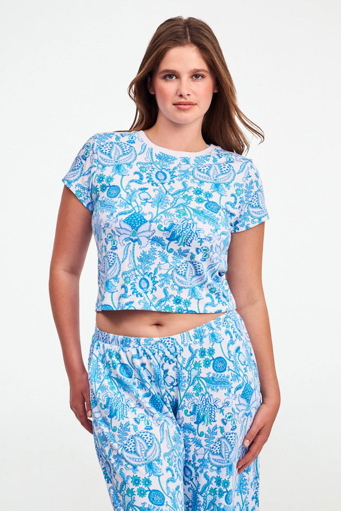 A woman with long brown hair wears the Roller Rabbit Amanda Baby Tee, featuring a blue and turquoise floral and sea-inspired print, standing against a plain white background.
