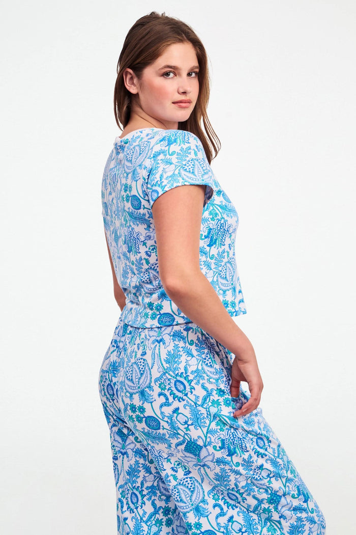 A woman with long brown hair wears the Roller Rabbit Amanda Baby Tee, featuring a blue and turquoise floral and sea-inspired print, standing against a plain white background.
