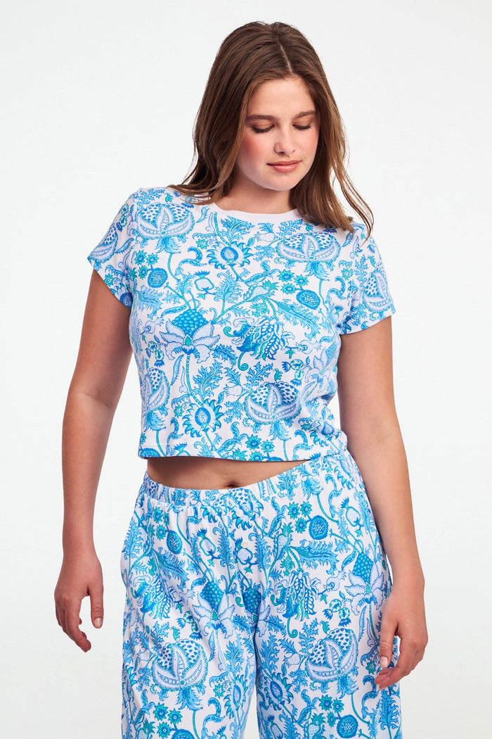 A woman with long brown hair wears the Roller Rabbit Amanda Baby Tee, featuring a blue and turquoise floral and sea-inspired print, standing against a plain white background.