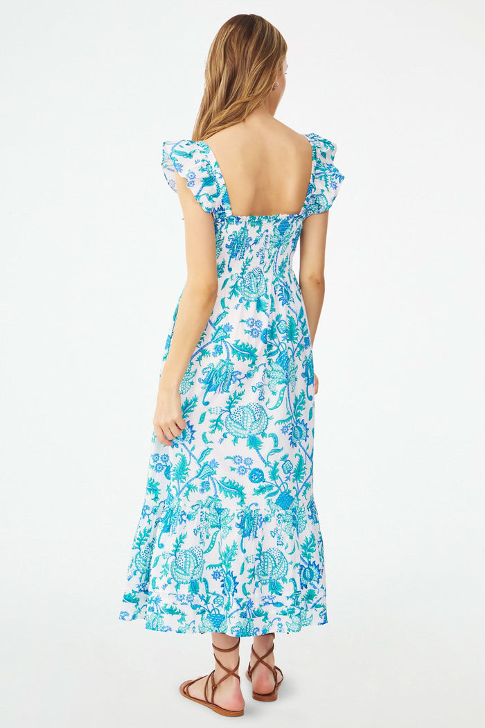 A woman models the Roller Rabbit Amanda Doe Dress—a blue and white cotton voile maxi with flutter sleeves, smocked bodice, and square neckline—paired with brown strappy sandals against a plain white background.