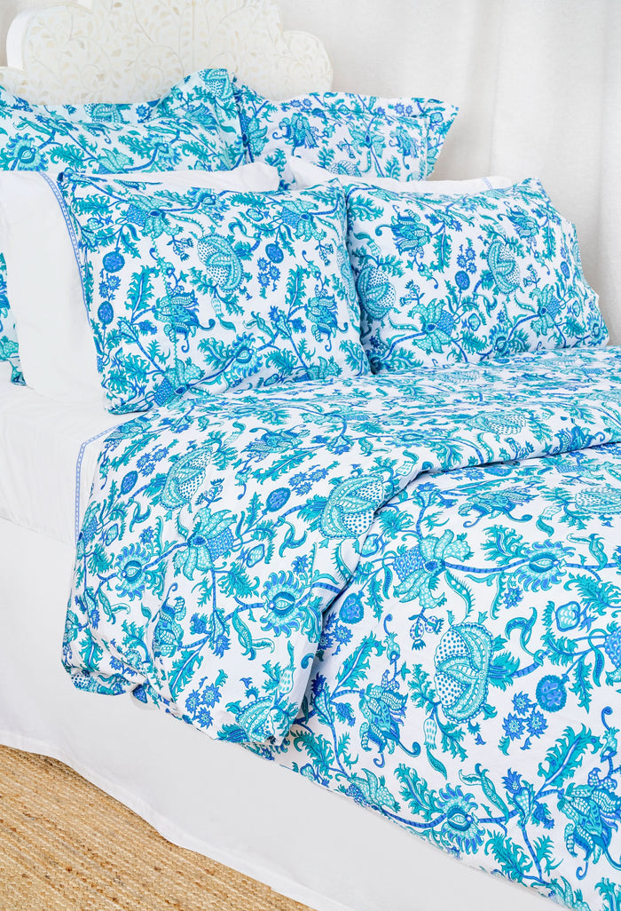 A neatly made bed features the Roller Rabbit Amanda Duvet Cover with blue and turquoise botanical print, matching pillow shams, white sheets, a white headboard and curtain, and a woven rug partly visible on the floor.