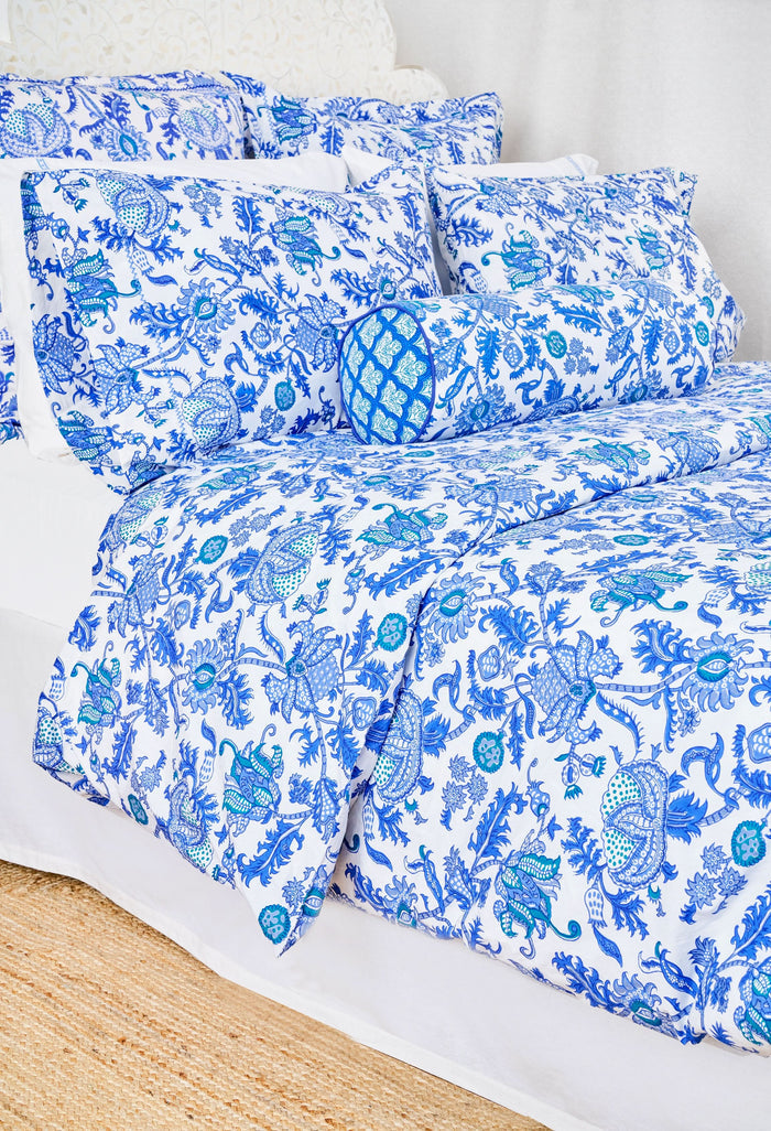 A neatly made bed features the Amanda Duvet Cover by Roller Rabbit in a white and blue botanical print, with matching pillows and a cylindrical bolster, all set against a white headboard and light background.