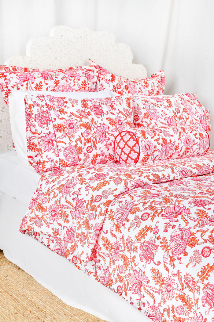 A bed featuring the Roller Rabbit Amanda Duvet Cover, a 300 thread count cotton piece in vibrant pink and orange botanical print, shown with a white carved headboard and white curtains in the background.