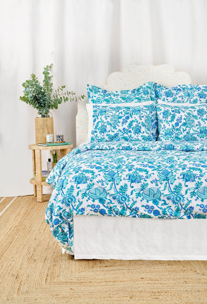 A neatly made bed features the Roller Rabbit Amanda Duvet Cover with blue and turquoise botanical print, matching pillow shams, white sheets, a white headboard and curtain, and a woven rug partly visible on the floor.