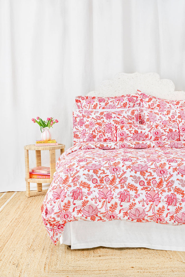 A bed featuring the Roller Rabbit Amanda Duvet Cover, a 300 thread count cotton piece in vibrant pink and orange botanical print, shown with a white carved headboard and white curtains in the background.