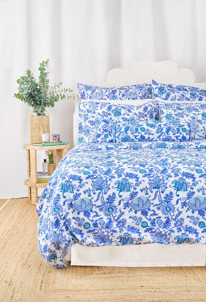 A neatly made bed features the Amanda Duvet Cover by Roller Rabbit in a white and blue botanical print, with matching pillows and a cylindrical bolster, all set against a white headboard and light background.