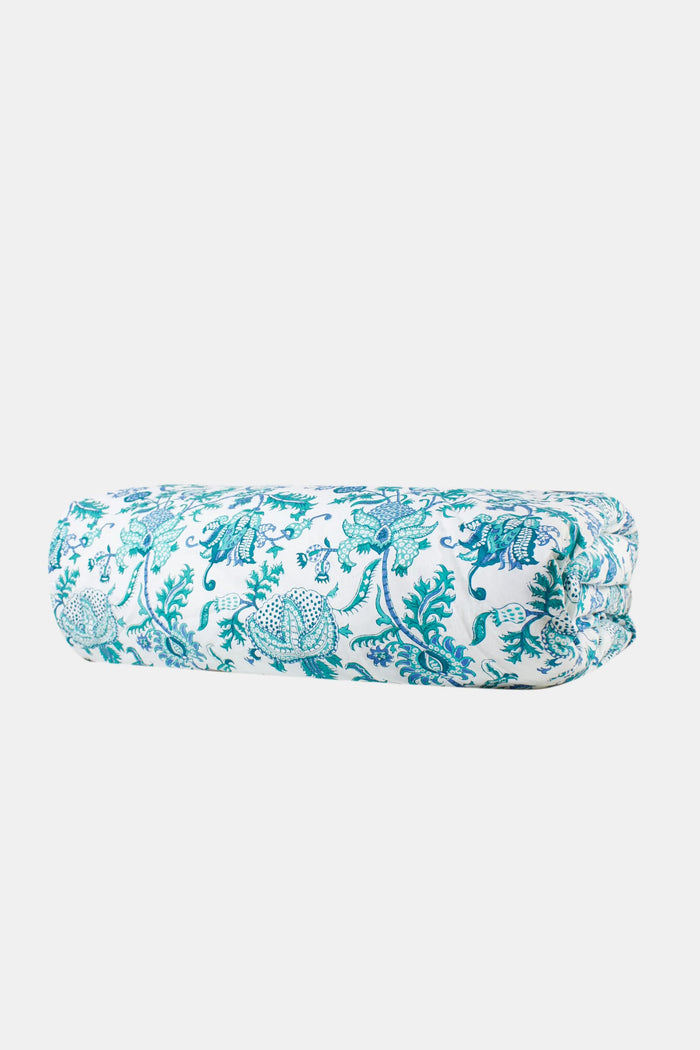 A neatly made bed features the Roller Rabbit Amanda Duvet Cover with blue and turquoise botanical print, matching pillow shams, white sheets, a white headboard and curtain, and a woven rug partly visible on the floor.