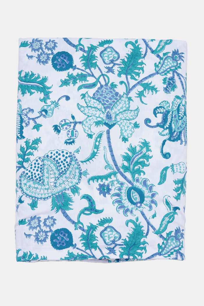 A neatly made bed features the Roller Rabbit Amanda Duvet Cover with blue and turquoise botanical print, matching pillow shams, white sheets, a white headboard and curtain, and a woven rug partly visible on the floor.