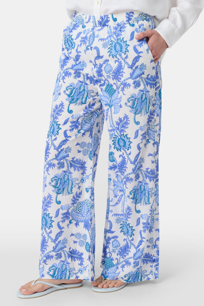 A person wears Roller Rabbit’s Amanda Kaylee Pant—white wide-leg lounge pants with a blue Amanda print and smocked waist—paired with a white shirt and flip-flops. Only the lower half of their body is visible, hand in one pocket.