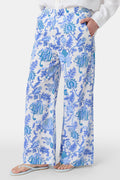 A person wears Roller Rabbit’s Amanda Kaylee Pant—white wide-leg lounge pants with a blue Amanda print and smocked waist—paired with a white shirt and flip-flops. Only the lower half of their body is visible, hand in one pocket.