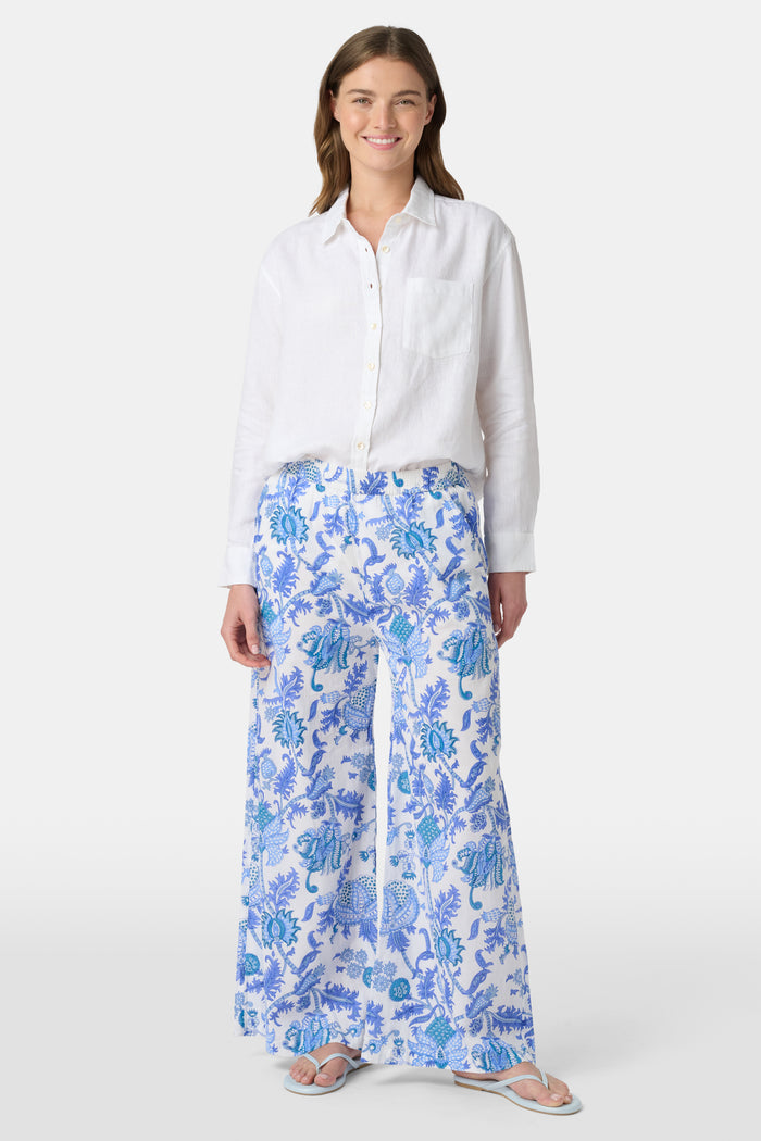 A person wears Roller Rabbit’s Amanda Kaylee Pant—white wide-leg lounge pants with a blue Amanda print and smocked waist—paired with a white shirt and flip-flops. Only the lower half of their body is visible, hand in one pocket.