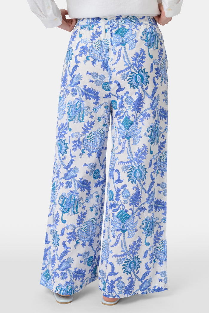 A person wears Roller Rabbit’s Amanda Kaylee Pant—white wide-leg lounge pants with a blue Amanda print and smocked waist—paired with a white shirt and flip-flops. Only the lower half of their body is visible, hand in one pocket.