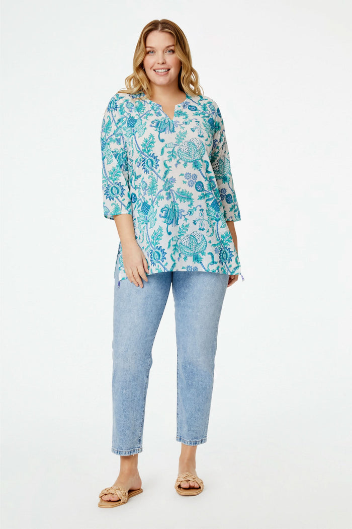 A smiling woman wears the Amanda Kurta by Roller Rabbit, a white piece adorned with blue and teal floral prints and hand-beaded details. She styles it with layered necklaces and bracelets against a plain, bright background.