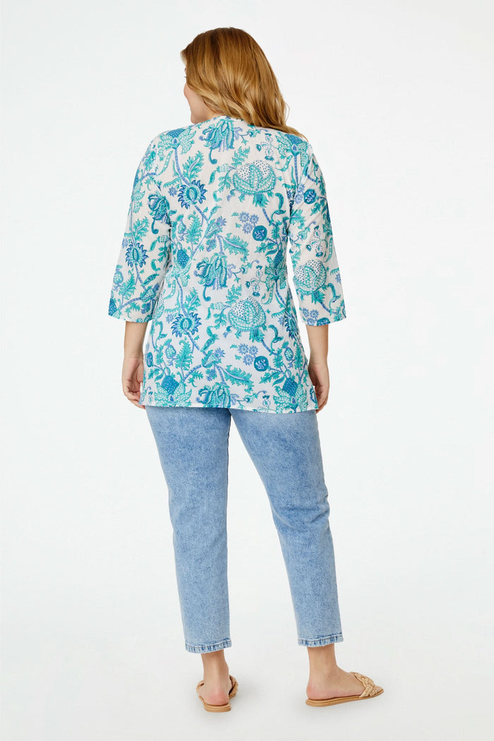 A smiling woman wears the Amanda Kurta by Roller Rabbit, a white piece adorned with blue and teal floral prints and hand-beaded details. She styles it with layered necklaces and bracelets against a plain, bright background.