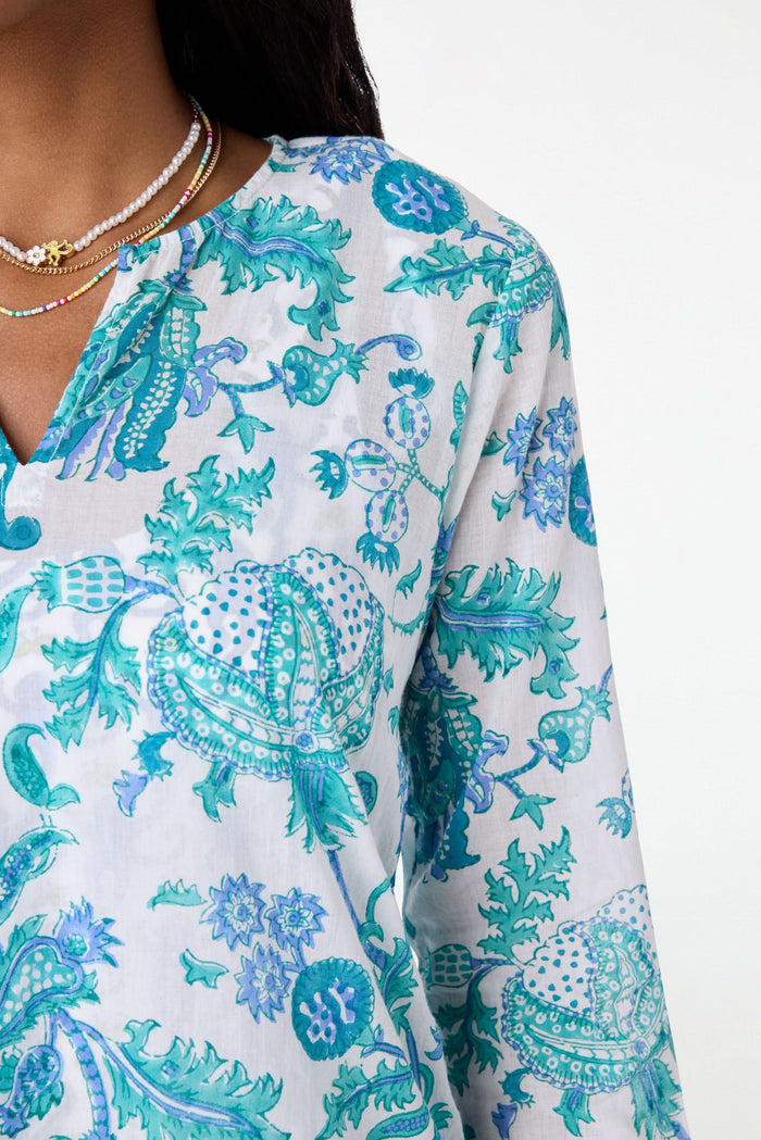 A smiling woman wears the Amanda Kurta by Roller Rabbit, a white piece adorned with blue and teal floral prints and hand-beaded details. She styles it with layered necklaces and bracelets against a plain, bright background.