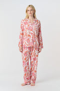 A woman with long blonde hair stands barefoot against a plain background, modeling the Amanda Loungewear by Roller Rabbit—an elegant women's pajama set featuring a white base with red and pink floral prints.
