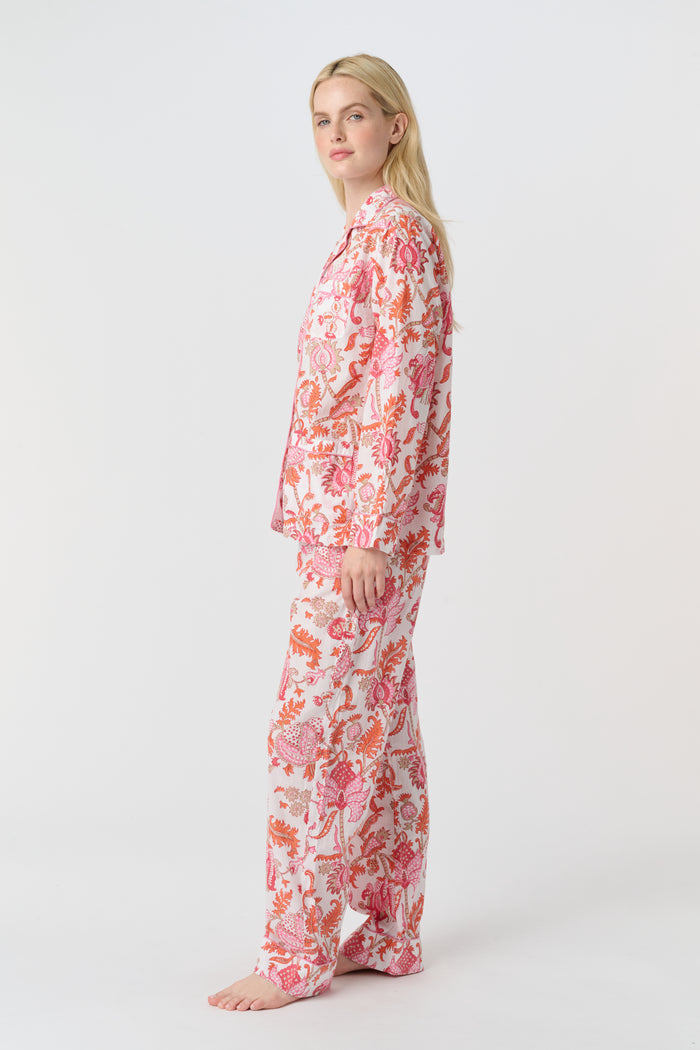 A woman with long blonde hair stands barefoot against a plain background, modeling the Amanda Loungewear by Roller Rabbit—an elegant women's pajama set featuring a white base with red and pink floral prints.