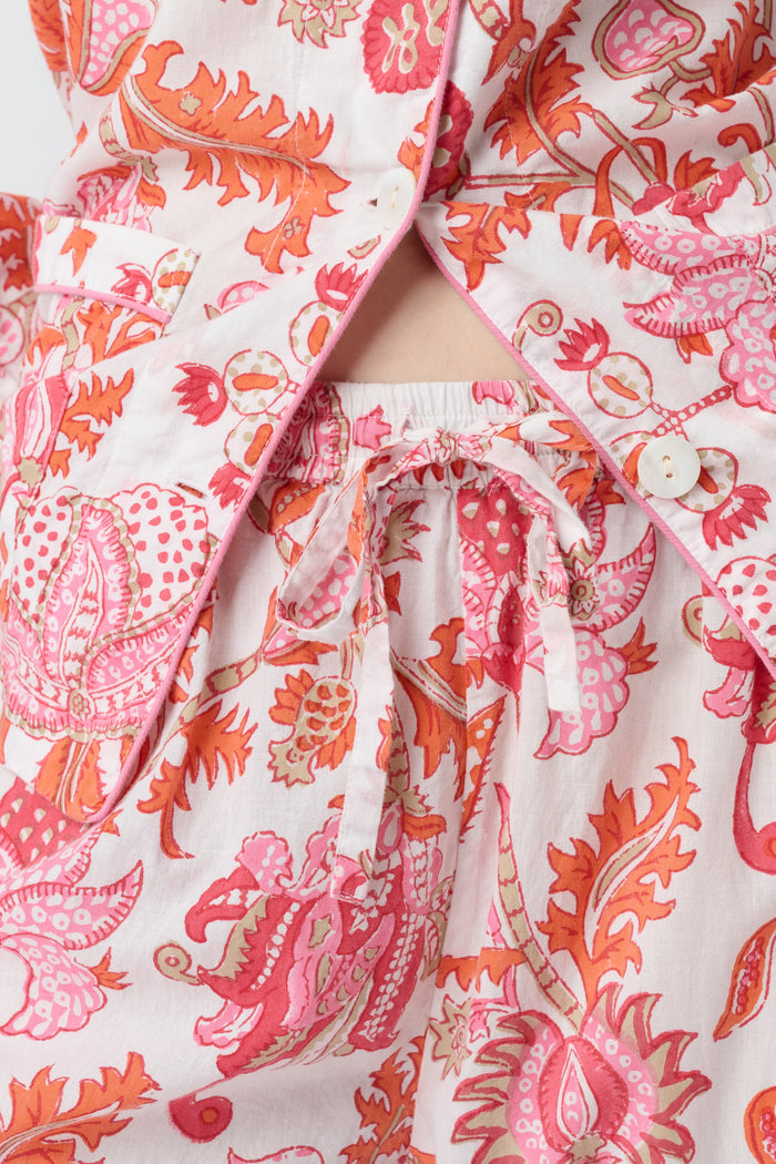 A woman with long blonde hair stands barefoot against a plain background, modeling the Amanda Loungewear by Roller Rabbit—an elegant women's pajama set featuring a white base with red and pink floral prints.