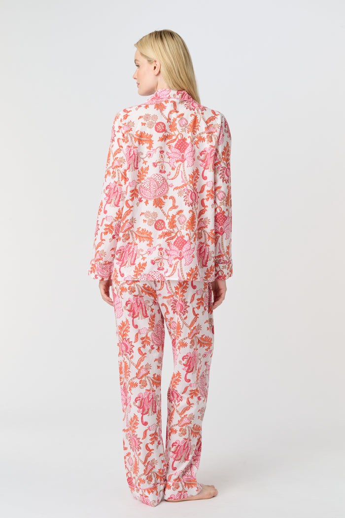 A woman with long blonde hair stands barefoot against a plain background, modeling the Amanda Loungewear by Roller Rabbit—an elegant women's pajama set featuring a white base with red and pink floral prints.