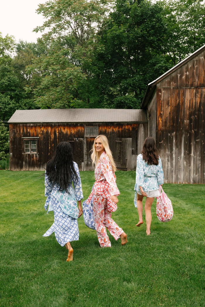 A woman with long blonde hair stands barefoot against a plain background, modeling the Amanda Loungewear by Roller Rabbit—an elegant women's pajama set featuring a white base with red and pink floral prints.