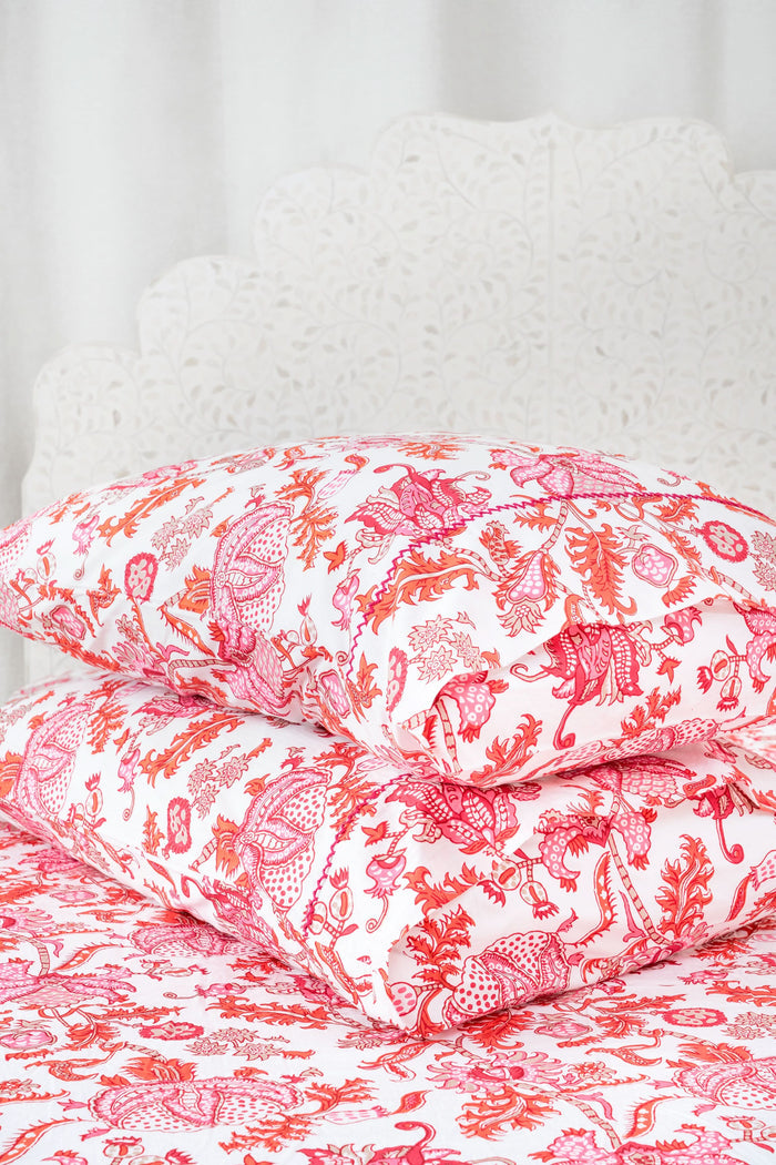 Two stacked pillows on a bed showcase the Roller Rabbit Amanda Pillowcase Set, featuring a vibrant red and pink floral and animal print on soft 300 thread count cotton, set against a white decorative headboard.