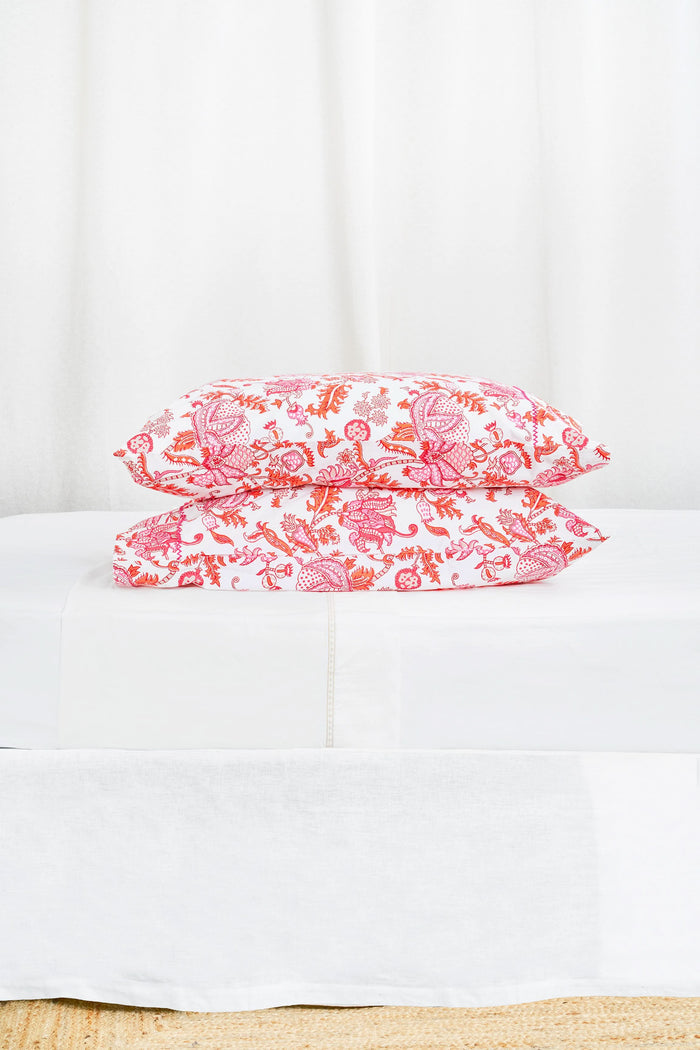 Two stacked pillows on a bed showcase the Roller Rabbit Amanda Pillowcase Set, featuring a vibrant red and pink floral and animal print on soft 300 thread count cotton, set against a white decorative headboard.