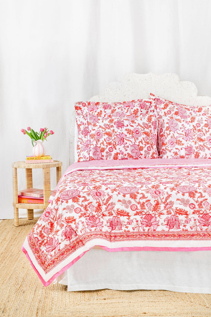 The Amanda Quilt by Roller Rabbit, featuring a white and pink floral cotton design with matching pillows, is displayed against a white backdrop next to a wooden side table holding pink tulips in a vase and stacked books.
