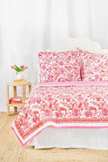 The Amanda Quilt by Roller Rabbit, featuring a white and pink floral cotton design with matching pillows, is displayed against a white backdrop next to a wooden side table holding pink tulips in a vase and stacked books.