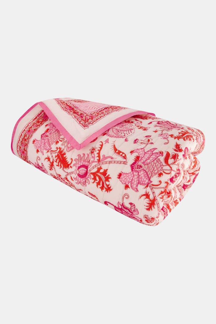 The Amanda Quilt by Roller Rabbit, featuring a white and pink floral cotton design with matching pillows, is displayed against a white backdrop next to a wooden side table holding pink tulips in a vase and stacked books.