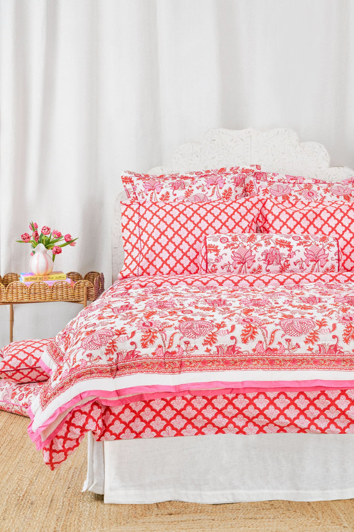 The Amanda Quilt by Roller Rabbit, featuring a white and pink floral cotton design with matching pillows, is displayed against a white backdrop next to a wooden side table holding pink tulips in a vase and stacked books.