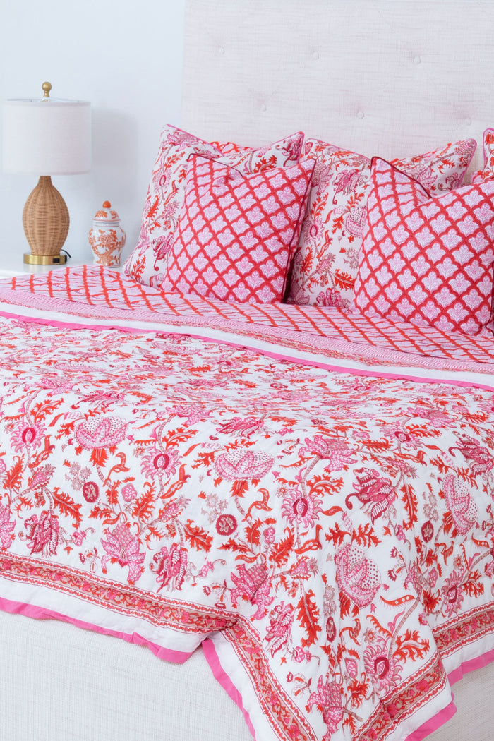 The Amanda Quilt by Roller Rabbit, featuring a white and pink floral cotton design with matching pillows, is displayed against a white backdrop next to a wooden side table holding pink tulips in a vase and stacked books.