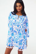 A woman with long dark hair wears the Amanda Serafina Tunic by Roller Rabbit, a loose, long-sleeved white top with a blue floral pattern and white tassels at the hem, standing against a plain light background.