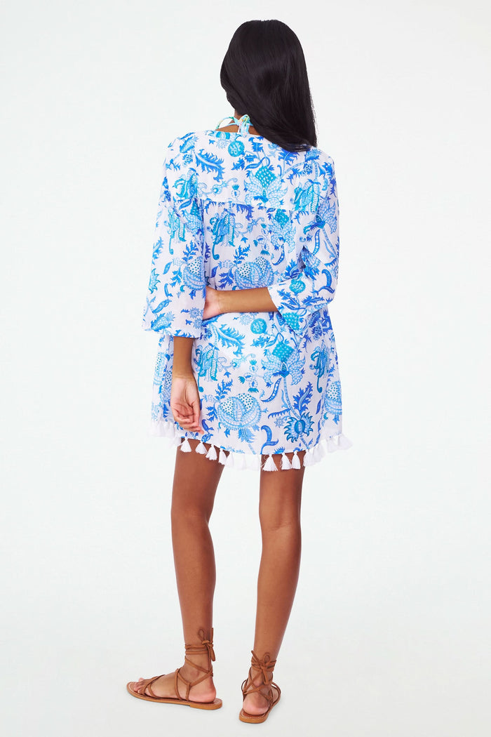 A woman with long dark hair wears the Amanda Serafina Tunic by Roller Rabbit, a loose, long-sleeved white top with a blue floral pattern and white tassels at the hem, standing against a plain light background.