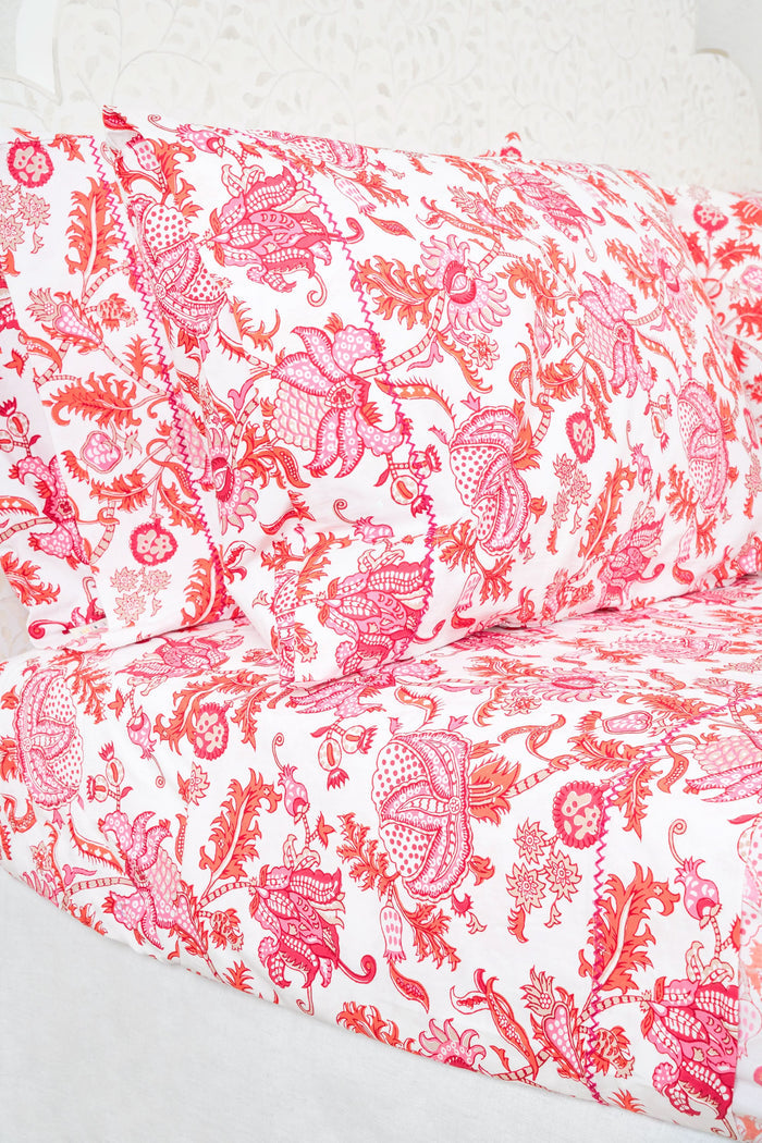 The bed displays the Amanda Sheet Set by Roller Rabbit, featuring white and pink botanical print sheets and pillowcases with intricate red and pink designs. A white headboard with subtle decorative detailing is visible in the background.