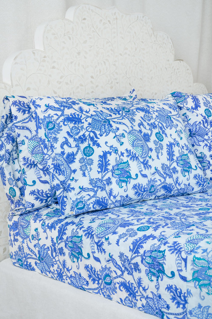 A bed dressed in the Amanda Sheet Set by Roller Rabbit, featuring white and blue botanical prints with intricate designs, is set against a white ornate headboard with a scalloped edge.