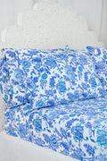 A bed dressed in the Amanda Sheet Set by Roller Rabbit, featuring white and blue botanical prints with intricate designs, is set against a white ornate headboard with a scalloped edge.