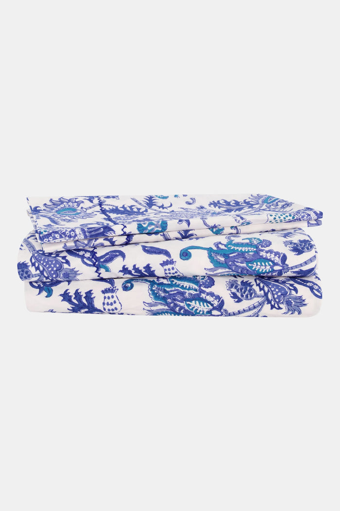 A bed dressed in the Amanda Sheet Set by Roller Rabbit, featuring white and blue botanical prints with intricate designs, is set against a white ornate headboard with a scalloped edge.