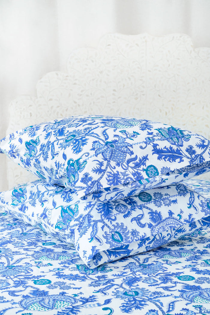 A bed dressed in the Amanda Sheet Set by Roller Rabbit, featuring white and blue botanical prints with intricate designs, is set against a white ornate headboard with a scalloped edge.