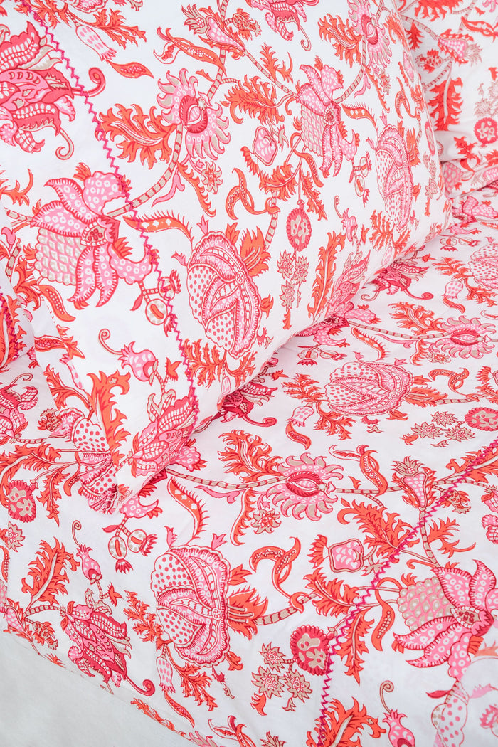 The bed displays the Amanda Sheet Set by Roller Rabbit, featuring white and pink botanical print sheets and pillowcases with intricate red and pink designs. A white headboard with subtle decorative detailing is visible in the background.