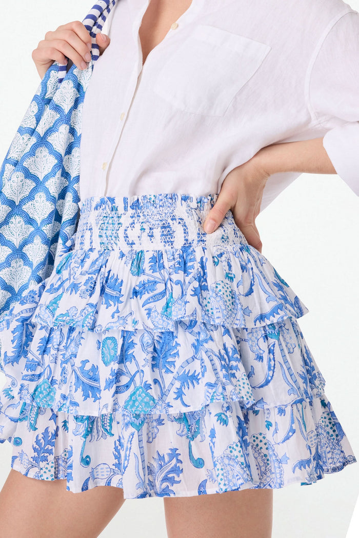 A person wearing a white button-up shirt and the Amanda Siggy Skirt by Roller Rabbit, featuring a blue print and smocked waist, holding a matching blue-and-white patterned bag.