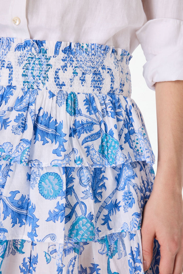 A person wearing a white button-up shirt and the Amanda Siggy Skirt by Roller Rabbit, featuring a blue print and smocked waist, holding a matching blue-and-white patterned bag.